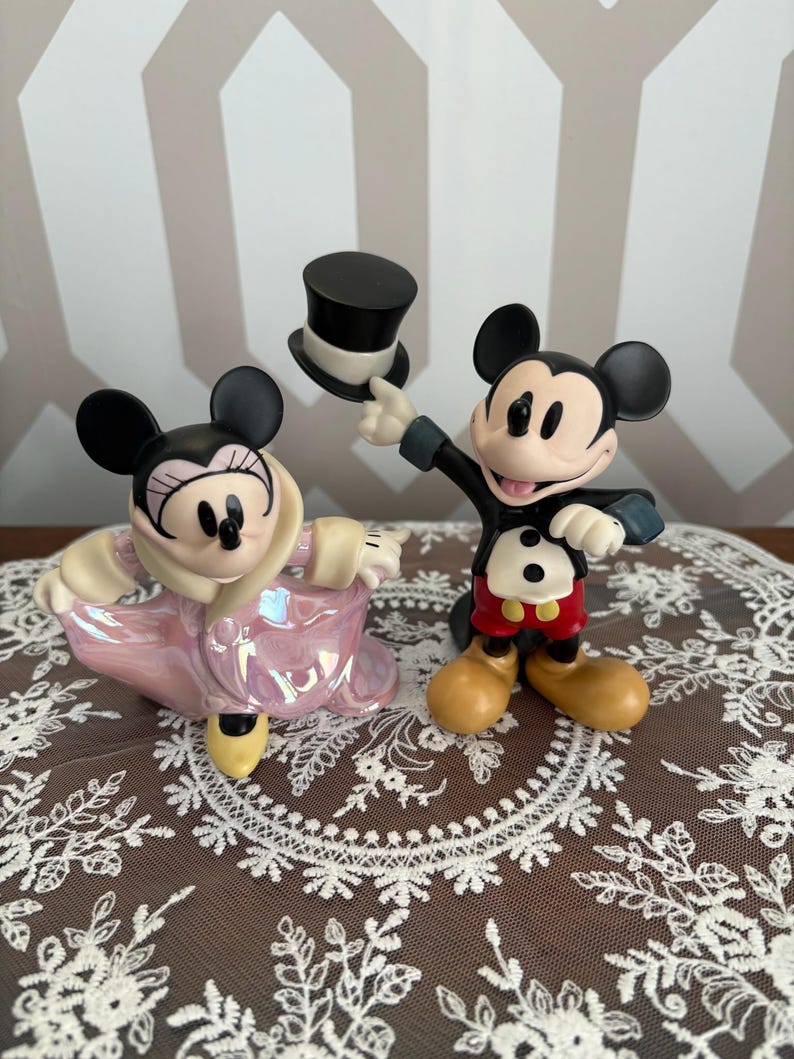 May include: Two ceramic figurines of Mickey and Minnie Mouse. Mickey is in a tuxedo, holding a black top hat. Minnie is wearing a pink iridescent coat. Both have black ears and are smiling.