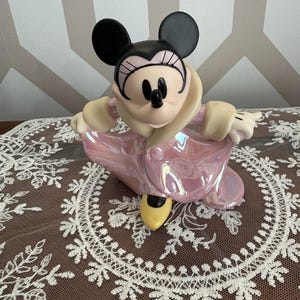 May include: A Minnie Mouse figurine in a pink iridescent coat with a cream-colored collar and black ears. The figurine has a black nose and eyes, and yellow shoes. The figurine is posed with arms outstretched.
