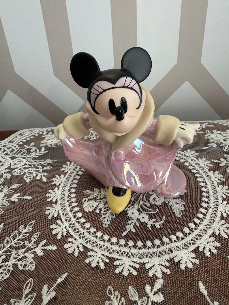 May include: A Minnie Mouse figurine in a pink iridescent coat with a cream-colored collar and black ears. The figurine has a black nose and eyes, and yellow shoes. The figurine is posed with arms outstretched.