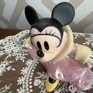 May include: A ceramic Minnie Mouse figurine in a seated pose. The figure has black ears, a black nose, and a pink iridescent dress. The figurine has yellow shoes and a cream-colored collar.