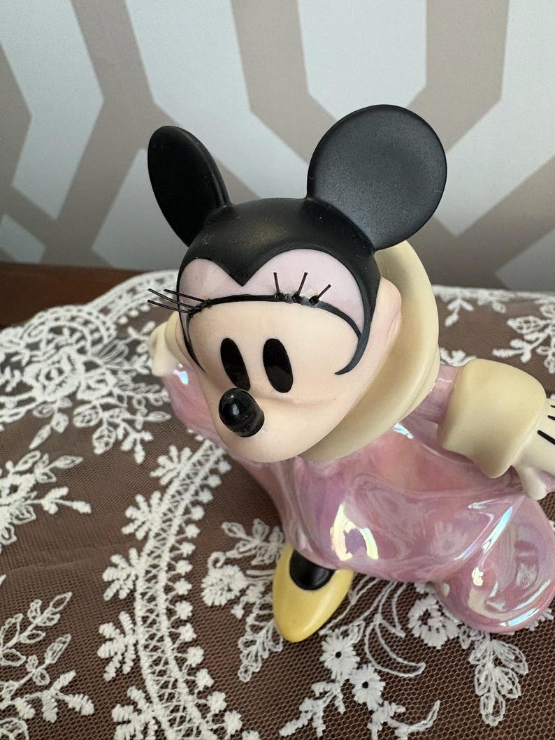 May include: A ceramic Minnie Mouse figurine in a seated pose. The figure has black ears, a black nose, and a pink iridescent dress. The figurine has yellow shoes and a cream-colored collar.