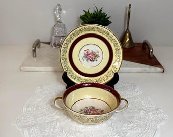 May include: A vintage teacup and saucer set. The set features a cream-colored base with a maroon border and gold trim. The center of the saucer and the inside of the teacup have a floral design. The teacup has two small handles.