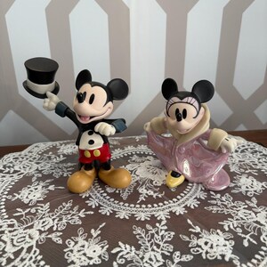 May include: Two Disney Mickey Mouse and Minnie Mouse figurines. Mickey is in a black tuxedo with a top hat, red shorts, and yellow shoes. Minnie is in a pink coat. Both have black ears and are smiling.