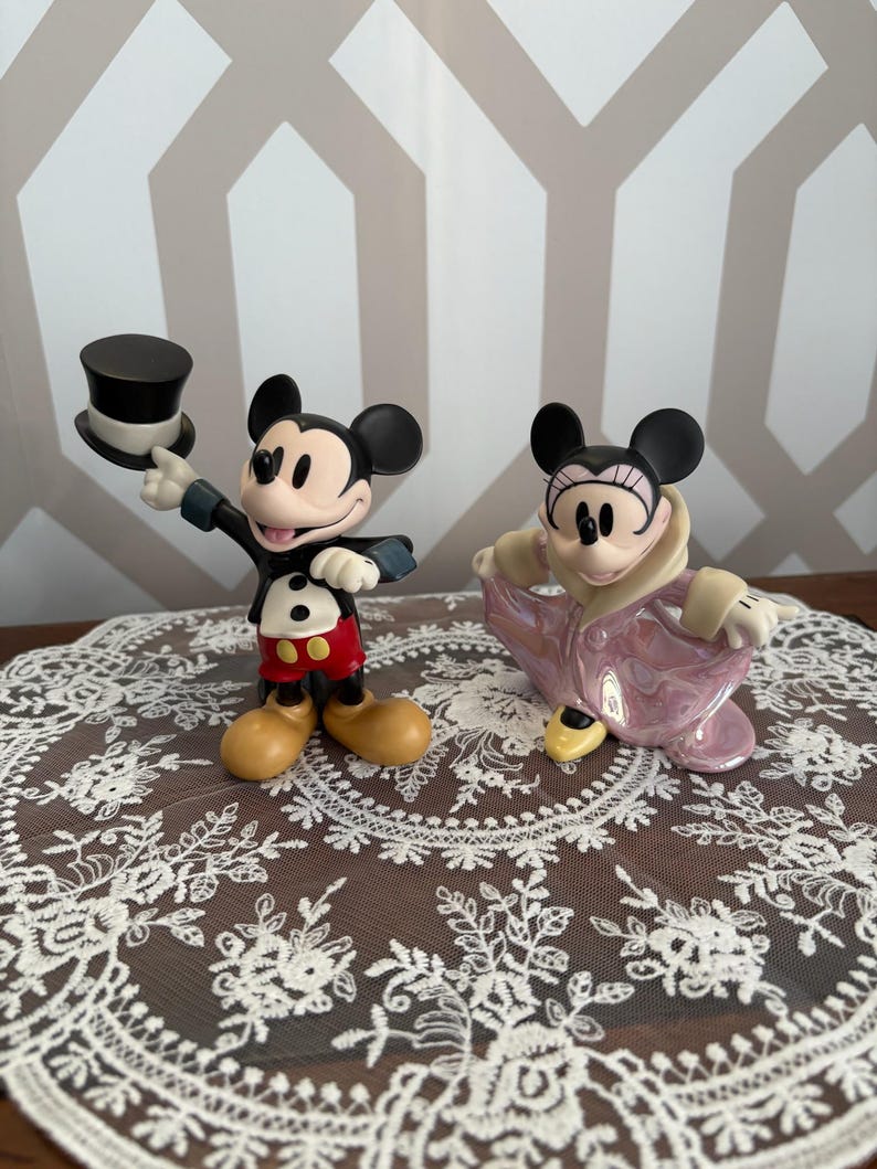 May include: Two Disney Mickey Mouse and Minnie Mouse figurines. Mickey is in a black tuxedo with a top hat, red shorts, and yellow shoes. Minnie is in a pink coat. Both have black ears and are smiling.