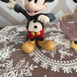 May include: A Mickey Mouse figurine in a classic tuxedo, holding a top hat. The figure has red shorts, yellow shoes, and a black jacket. The figurine is on a brown surface with white lace.