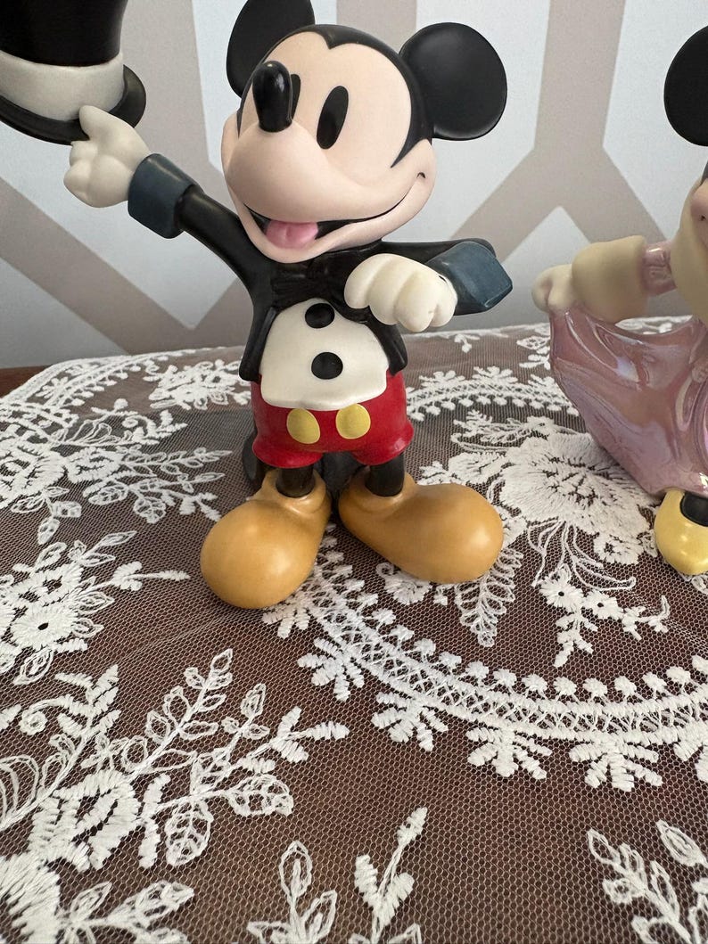 May include: A Mickey Mouse figurine in a classic tuxedo, holding a top hat. The figure has red shorts, yellow shoes, and a black jacket. The figurine is on a brown surface with white lace.