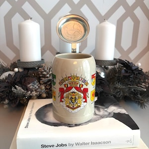 May include: A beige ceramic beer stein with a hinged metal lid. The stein features a detailed coat of arms design with multiple crests and a crown. The background includes white candles and a decorative garland.