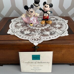 May include: A pair of Mickey and Minnie Mouse figurines, Mickey holding a top hat. Minnie wears a pink dress and coat. They are on a white lace doily atop a wooden box. A certificate of authenticity is in front of the box.