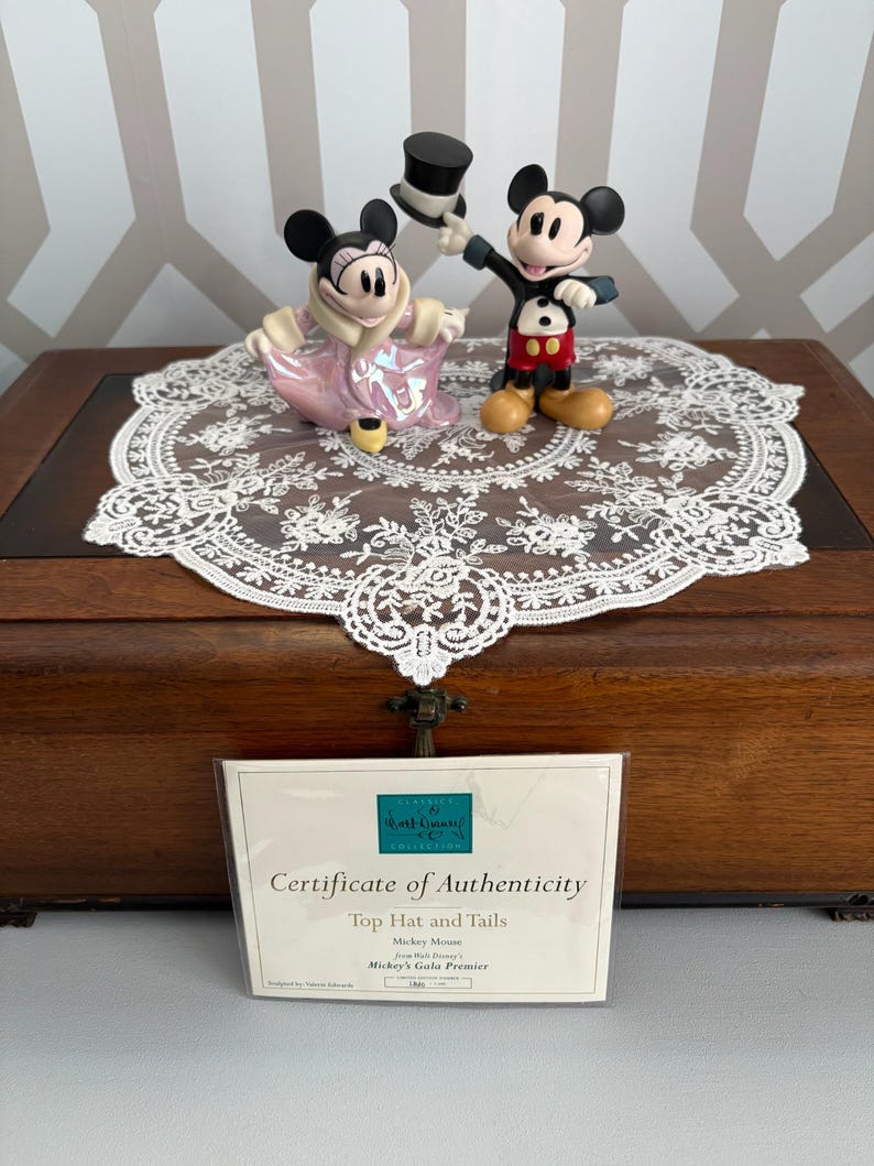 May include: A pair of Mickey and Minnie Mouse figurines, Mickey holding a top hat. Minnie wears a pink dress and coat. They are on a white lace doily atop a wooden box. A certificate of authenticity is in front of the box.