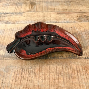 May include: A leaf-shaped ashtray with a glossy finish. The ceramic ashtray features a dark brown base with red and black accents. It has three grooves for holding cigarettes.