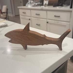 Könnte beinhalten: Eine Holz-Hai-Figur, hergestellt aus warmbraunem Holz. Der Hai hat eine Rückenflosse, einen detaillierten Schwanz und ein offenes Maul. Die Figur wird auf einer weißen Arbeitsplatte präsentiert, die die Handwerkskunst der Holzskulptur hervorhebt.