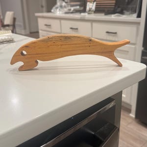 Puede incluir: Un abridor de botellas de madera con forma de pez. El abridor está hecho de madera de color claro con un acabado natural. Tiene un cuerpo curvado, una cola puntiaguda y una abertura en forma de boca para quitar las tapas. El pez mide unos 30 cm de largo.