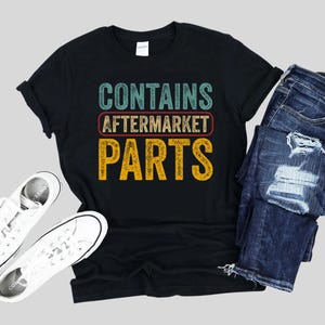 Könnte beinhalten: Schwarzes T-Shirt mit dem Text "CONTAINS AFTERMARKET PARTS" in Türkis, Rot und Gelb. Das Shirt wird mit einer blauen Jeans und weißen Turnschuhen kombiniert. Die Jeans hat Distressed-Details.