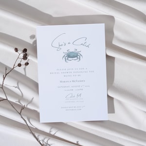May include: A white invitation card featuring the text "She's a Catch" and a blue-green crab illustration. The card is for a bridal shower, including the bride's name, date, time, and venue details.