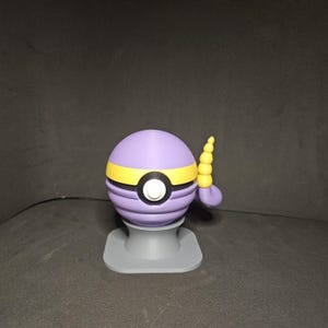 May include: A purple and yellow spherical figurine with a white center, black outline, and a small tail. The figurine is on a gray base. The design is reminiscent of a cartoon character.