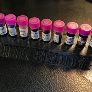 May include: A black plastic ink bottle holder with the words "INK BUDDY" printed on the front. The holder has ten slots for ink bottles. Nine bottles are filled with ink and have pink lids. The ink bottles are labelled with names such as "Muddy Swamp", "Muddy Wagon", "Muddy Crown", and "Grey Seas".
