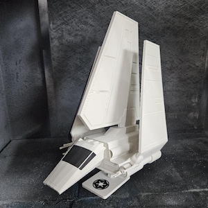 Könnte beinhalten: Ein weißes Modell eines Raumschiffs mit großen, abgewinkelten Flügeln und einem zentralen Rumpf. Das Modell verfügt über ein detailliertes Cockpit und das imperiale Symbol auf einer Basis. Detaillierte Nachbildung eines fiktiven Fahrzeugs.