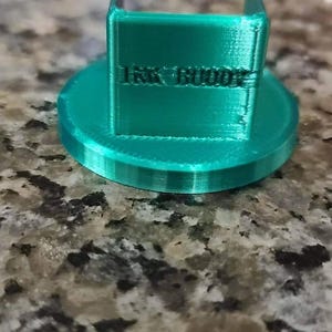 Op de afbeelding: Een 3D-geprint object van tealgroen met een vierkante basis en een rechthoekige bovenkant. Op de bovenkant staat de tekst "Ink Buddy" gedrukt.