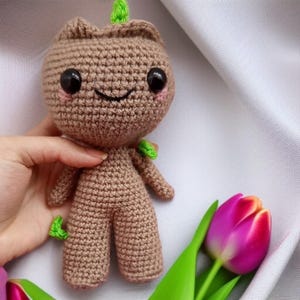 Pode incluir: Um personagem Groot de crochê marrom com uma parte superior verde frondosa e grandes olhos pretos. O personagem está sorrindo e tem um rubor rosado nas bochechas. O personagem está em pé sobre uma superfície branca.