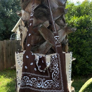 Puede incluir: Un bolso tote confeccionado en tela con un estampado de bandana de cachemira en marr&oacute;n y crema. El bolso incluye dos correas largas para el hombro y una textura acolchada con l&iacute;neas de costura verticales en toda la superficie exterior.