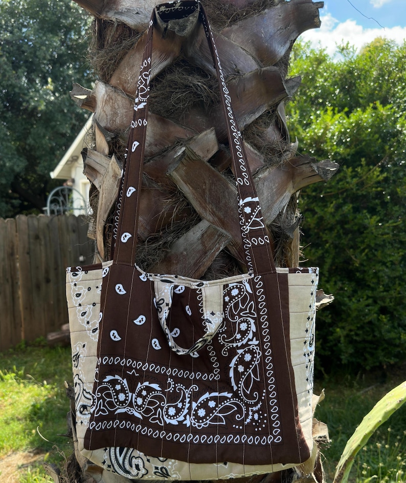 Puede incluir: Un bolso tote confeccionado en tela con un estampado de bandana de cachemira en marr&oacute;n y crema. El bolso incluye dos correas largas para el hombro y una textura acolchada con l&iacute;neas de costura verticales en toda la superficie exterior.