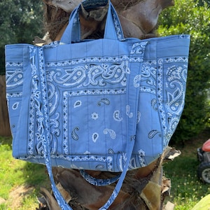 Puede incluir: Un bolso tote de color azul claro con un estampado cl&aacute;sico de pa&ntilde;uelo paisley en blanco y negro. El bolso tiene dos asas superiores y una correa larga para el hombro, con costuras acolchadas verticales en toda la tela y una base rectangular.