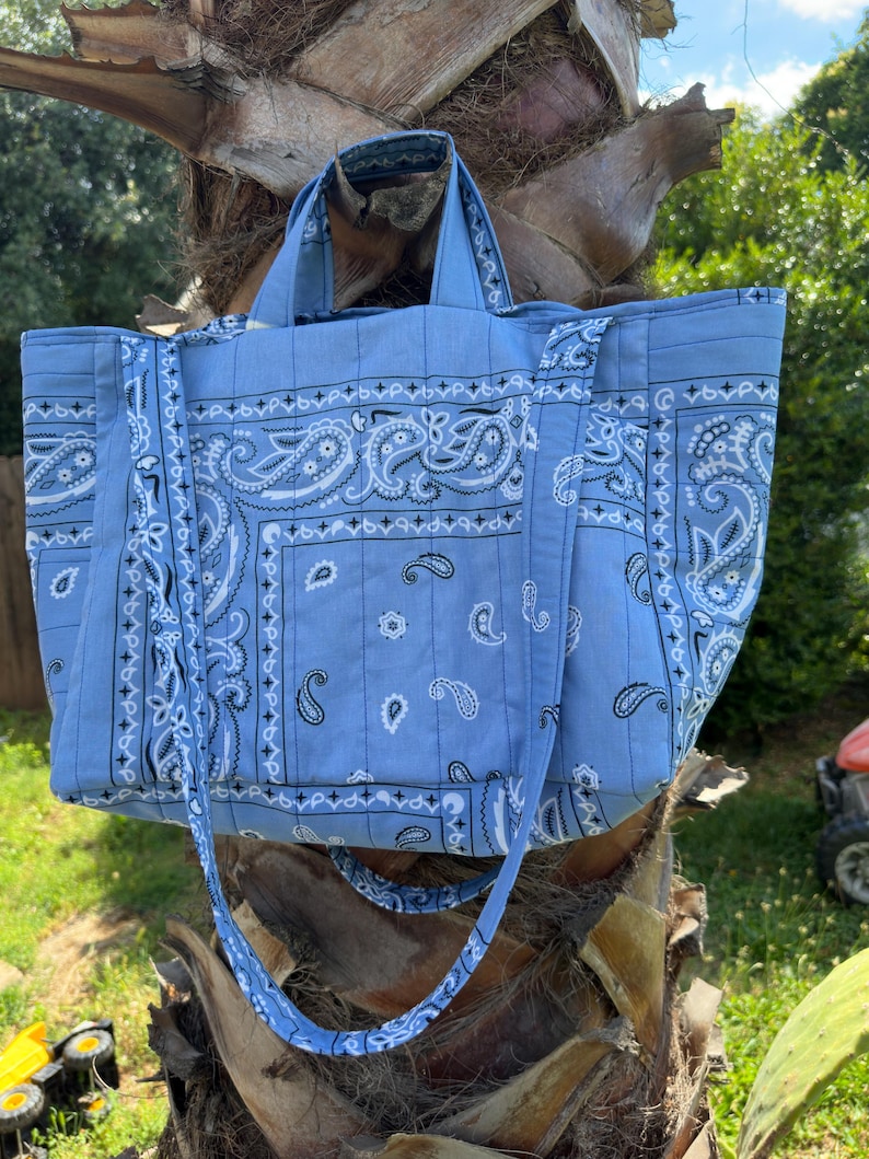Puede incluir: Un bolso tote de color azul claro con un estampado cl&aacute;sico de pa&ntilde;uelo paisley en blanco y negro. El bolso tiene dos asas superiores y una correa larga para el hombro, con costuras acolchadas verticales en toda la tela y una base rectangular.