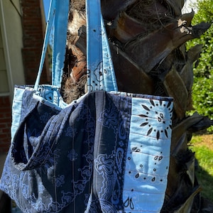 Puede incluir: Un bolso tote acolchado con un dise&ntilde;o de patchwork de telas estilo bandana en azul marino oscuro y azul claro. El bolso incluye dos correas largas para el hombro hechas de la misma tela azul claro estampada y costuras verticales.