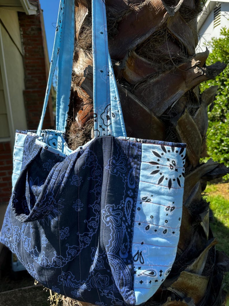 Puede incluir: Un bolso tote acolchado con un dise&ntilde;o de patchwork de telas estilo bandana en azul marino oscuro y azul claro. El bolso incluye dos correas largas para el hombro hechas de la misma tela azul claro estampada y costuras verticales.