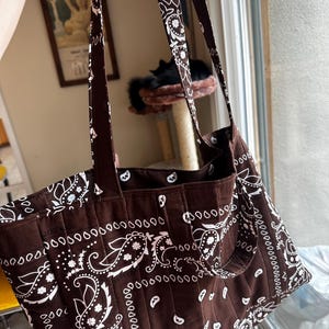 Puede incluir: Un bolso tote marr&oacute;n con un estampado de pa&ntilde;uelo paisley en blanco. El bolso est&aacute; confeccionado con costuras acolchadas verticales en todo el cuerpo e incluye dos correas largas para el hombro unidas al borde superior del compartimento.