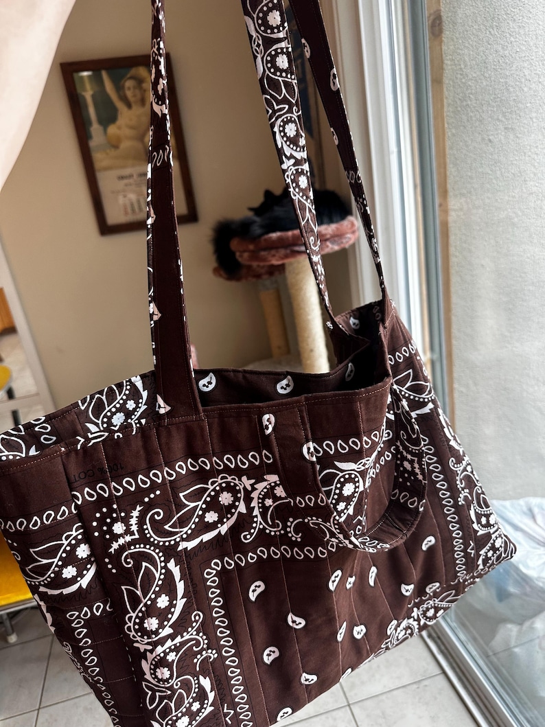 Puede incluir: Un bolso tote marr&oacute;n con un estampado de pa&ntilde;uelo paisley en blanco. El bolso est&aacute; confeccionado con costuras acolchadas verticales en todo el cuerpo e incluye dos correas largas para el hombro unidas al borde superior del compartimento.