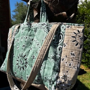 Puede incluir: Un bolso tote acolchado con un estampado de bandana en verde salvia y beige con motivos florales y de cachemira en negro. El bolso incluye dos asas superiores y una estructura rectangular con costuras verticales en los paneles de tela.