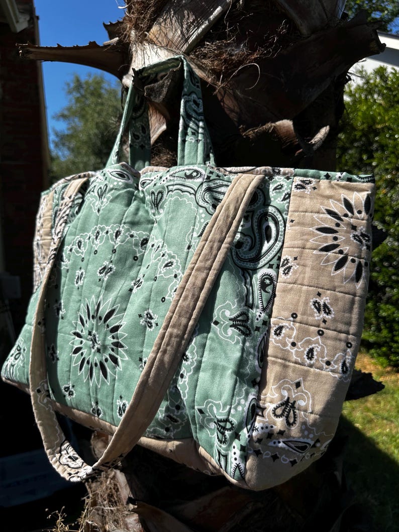 Puede incluir: Un bolso tote acolchado con un estampado de bandana en verde salvia y beige con motivos florales y de cachemira en negro. El bolso incluye dos asas superiores y una estructura rectangular con costuras verticales en los paneles de tela.