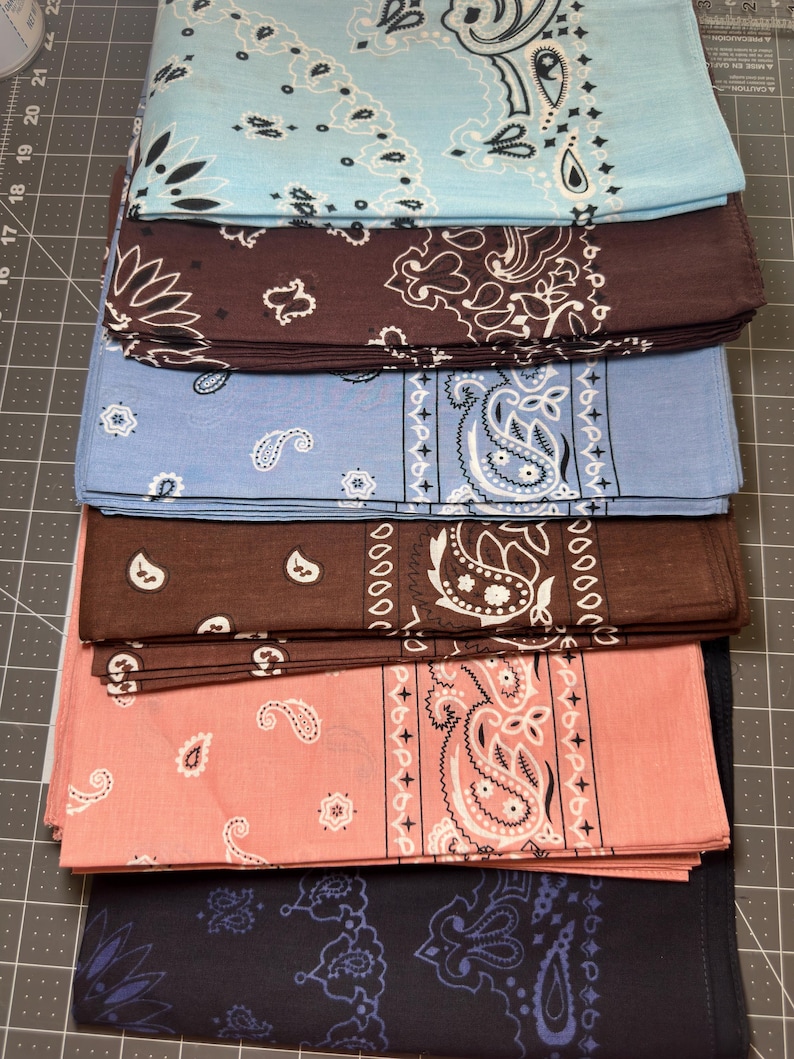 Puede incluir: Una pila de seis pa&ntilde;uelos cuadrados doblados con patrones de cachemira tradicionales. Los colores incluyen azul claro, marr&oacute;n oscuro, azul medio, marr&oacute;n chocolate, rosa salm&oacute;n y azul marino, dispuestos en una pila sobre una base de corte.