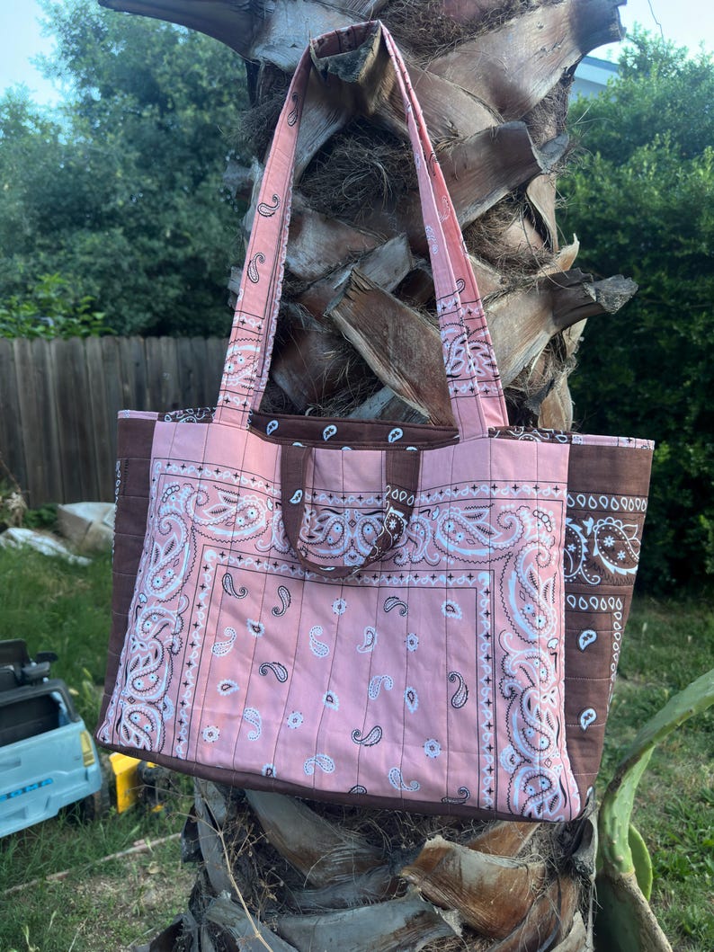Puede incluir: Un bolso tote acolchado con un estampado de cachemira tipo bandana en rosa y blanco en los paneles frontal y posterior, contrastado por paneles laterales y asas de color marr&oacute;n oscuro. Incluye dos correas largas y dos asas cortas.