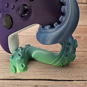 Pode incluir: Um controle Xbox roxo repousa sobre um suporte em forma de polvo impresso em 3D. O suporte transita do azul para o verde, com detalhes de tentáculos. O controle é de cor roxa escura.