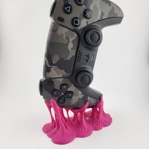Könnte beinhalten: Ein Videospiel-Controller mit grauem Camouflage-Muster und schwarzen Knöpfen und Akzenten, der auf einer hellrosa, strukturierten Basis ruht. Der Controller verfügt über ein Steuerkreuz, Aktionstasten und Analogsticks. Die Basis hat ein einzigartiges, geschmolzenes Design.