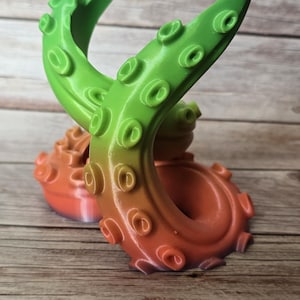 Może przedstawiać: 3D-drukowana rzeźba macki ośmiornicy w odcieniach zieleni i pomarańczy. Macka posiada przyssawki i efekt przejścia kolorów. Rzeźba jest prezentowana na drewnianej powierzchni.