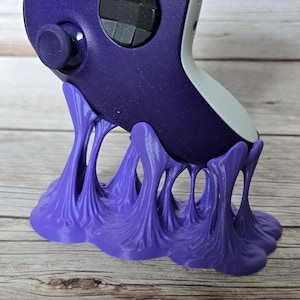 Può includere: Un controller per videogiochi viola è appoggiato su un supporto viola abbinato. Il supporto ha un design unico e fluido, simile a una macchia o a un liquido. Il controller è viola scuro con pulsanti neri e un retro bianco.