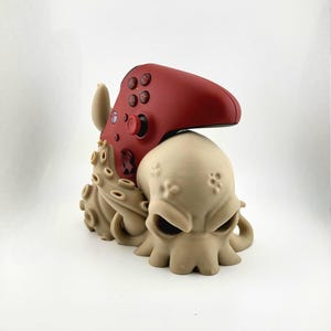 Könnte beinhalten: Ein beigefarbener, 3D-gedruckter Controller-Ständer im Oktopus-Design mit einem roten Controller obenauf. Der Ständer hat ein detailliertes Oktopus-Design mit Tentakeln und einem schädelartigen Kopf.