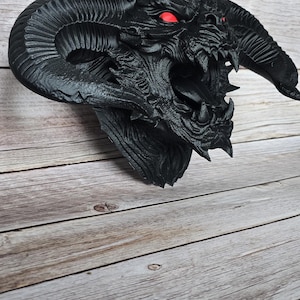 Op de afbeelding: Een gedetailleerde zwarte drakenhoofdsculptuur met prominente hoorns en scherpe tanden. De ogen van de draak zijn opvallend rood. De sculptuur is gemonteerd op een houten oppervlak.