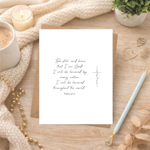 May include: A white rectangular card featuring black cursive text that reads: Be still, and know that I am God! I will be honored by every nation. I will be honored throughout the world. Psalms 46:10. A thin black cross with vine details is on the right.