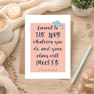 May include: A greeting card with the text "Commit to THE LORD whatever you do, and your plans will SUCCEED. Proverbs 16:3" in navy blue on a pink background. A blue graduation cap is at the top. The card is on a wooden surface.