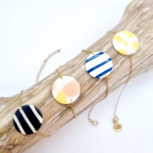 Peut inclure: Quatre colliers avec des pendentifs circulaires. Les pendentifs présentent divers motifs : rayures bleu marine et blanches, rayures bleues et blanches, pois jaunes et un mélange de formes roses et jaunes. Les pendentifs sont reliés par des chaînes dorées.