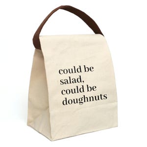 Pode incluir: Uma bolsa de almoço de lona branca com alças marrons. A bolsa tem o texto "could be salad, could be doughnuts" impresso na frente.