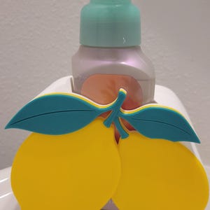 Può includere: Un dispenser di sapone decorativo con un design a limone giallo e foglie turchesi. Il dispenser ha una pompa verde chiaro e una bottiglia perlata. Un accessorio allegro e luminoso per il bagno o la cucina.