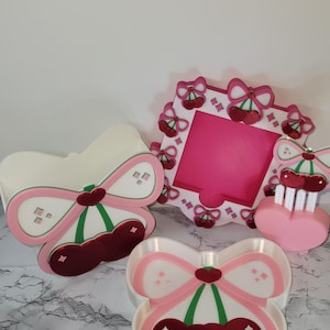 May include: A collection of decorative items with a cherry theme. Includes a picture frame, a heart-shaped item, and several trays. The items are white with pink and red accents, featuring cherry and bow designs. The overall aesthetic is cute and playful.