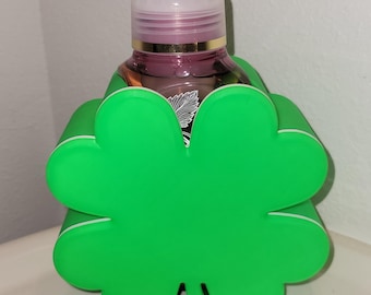 Könnte beinhalten: Ein grüner, vierblättriger Kleeblatt-Seifenspenderhalter mit weißem Rand. Der Halter umschließt eine transparente Seifenflasche mit rosa Flüssigkeit. Der Spender steht auf einer weißen Oberfläche.
