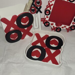 Puede incluir: Colección de decoración temática de San Valentín con diseños XOXO rojos y negros. Incluye un marco de fotos, un recipiente y detalles decorativos. Los artículos tienen una base blanca con X rojas, O negras y detalles de corazón.