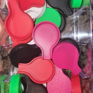 May include: A collection of colorful, 3D-printed objects in a clear plastic container. The objects are various shades of red, pink, green, and black. The shapes appear to be molds or cutters, with a rounded top and a handle.